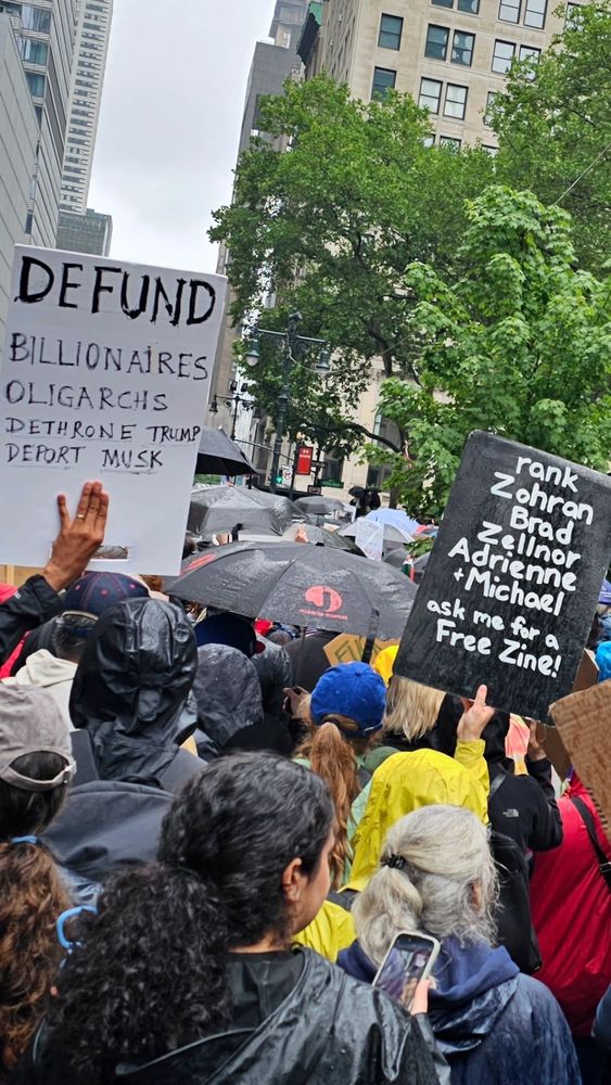 Backs of protesters with one sign that reads Defund billionaires, oligarchs,dethrone Trump, deport Musk” and another sign reading “rank Zohran, Brad, Zellnor, Adrienne and Michael, ask me for a free zine!”