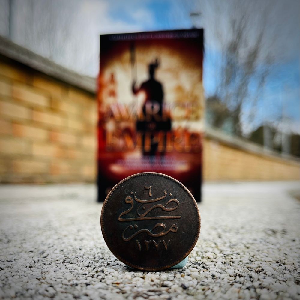 Nineteenth-century Egyptian 20 para coin in front of a copy of Avarice of Empire. 