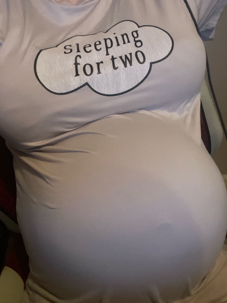 Mystery sitting in a chair wearing a pink nightgown that says “sleeping for two.” Her bloated belly clearly curves out from the tight garment, her belly button protruding under the cloth.