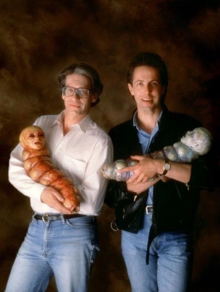 David Cronenberg and Clive Barker, dressed like '90s sitcom dads, holding worm alien babies from Nightbreed.
