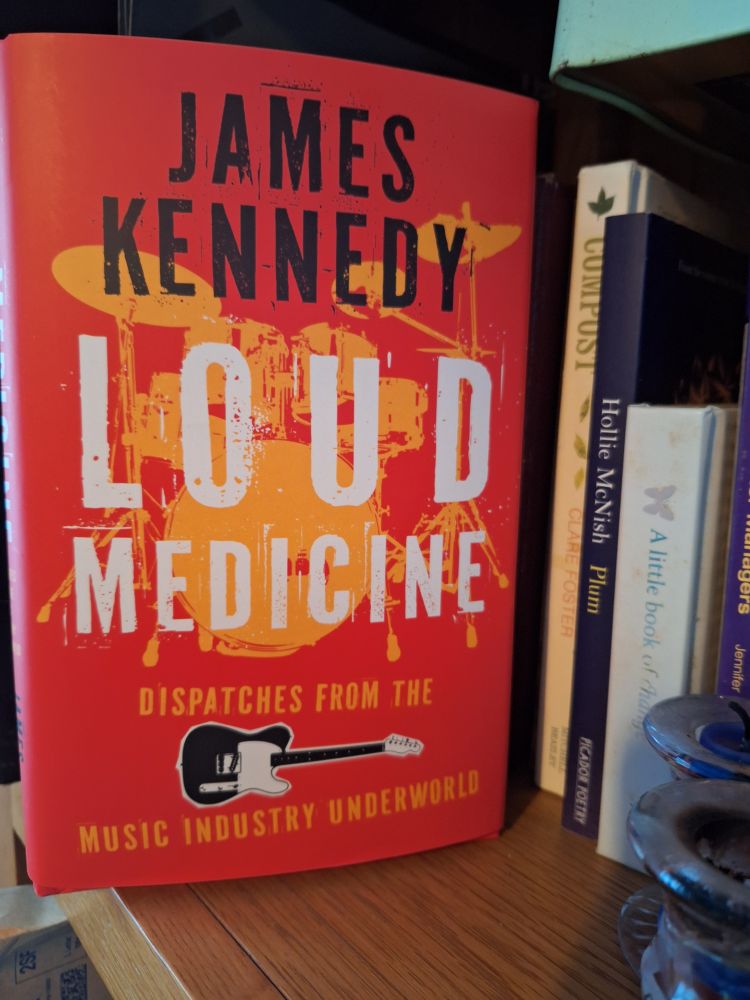Loud Medicine by James Kennedy tries to find room on an ill kempt book shelf. Book has vivid orange colouring and authors name in black lettering with title in big white letters. Picture of guitar near bottom of cover.