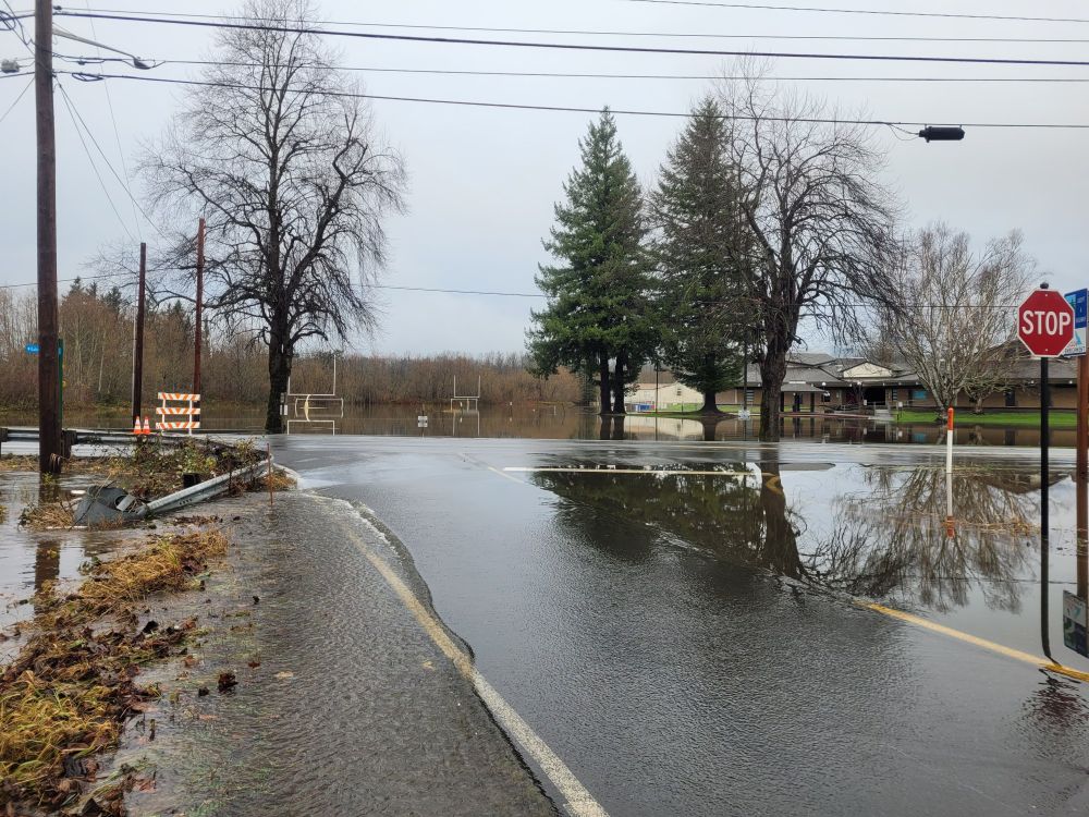 Looking out at SR-202 from Stone Quarry Road SE. The water retention ditch that runs along 202 has overtopped its bank. Water flows in a steady stream across Stone Quarry Road into the wetland on the other side, which is also full. Highway 202 is a bit higher and is above the water level. On the other side of 202 is Snoqualmie Middle School, the football field and drop-off roundabout still underwater.