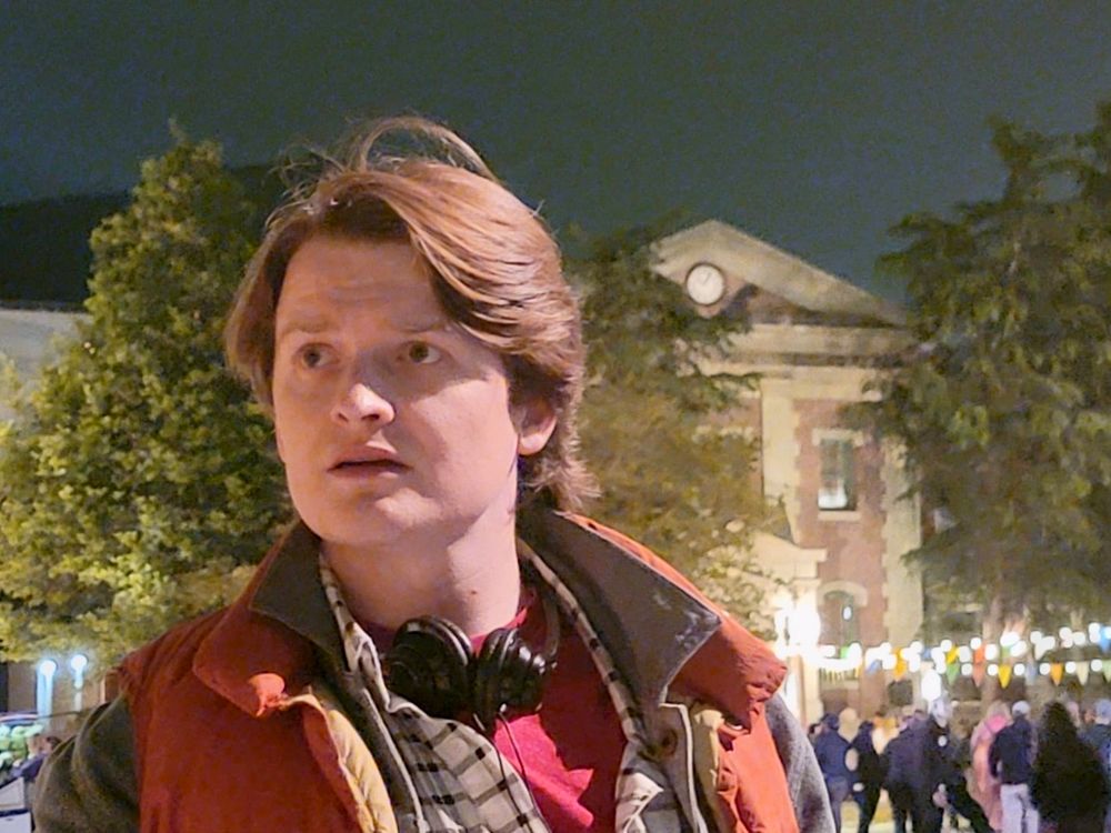 A photograph taken on the evening of 2025-05-04 in the Universal Studios Hollywood backlot, depicting a young man, myself, dressed in cosplay as Marty McFly from the 1985 film, “Back to the Future”. In the photo, I stand in the Courthouse Square set, with the clock tower standing some hundred yards behind me. The clock tower’s appearance has been modified since the production of “Back to the Future”, and now only bears a passing resemblance to the one seen in the film.