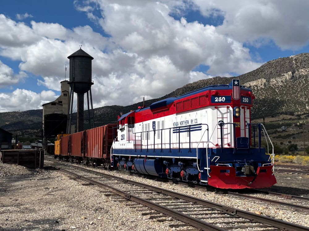 A photograph, taken 2025-09-20, featuring a locomotive and train sitting in a railroad yard in the Nevada High Desert. The locomotive, an SD-9R numbered 250, belongs to the Nevada Northern Railway Museum of Ely, Nevada. 250 wears a special red-white-and-blue livery, to commemorate the upcoming American Semiquincentennial; hence her number, “250”. Behind the locomotive are three brick-red ballast hopper cars, and an orange-yellow caboose. Further in the background are the coal and water towers of the East Ely yard complex, followed behind by a sagebrush-covered mountainside rising up from the valley floor.