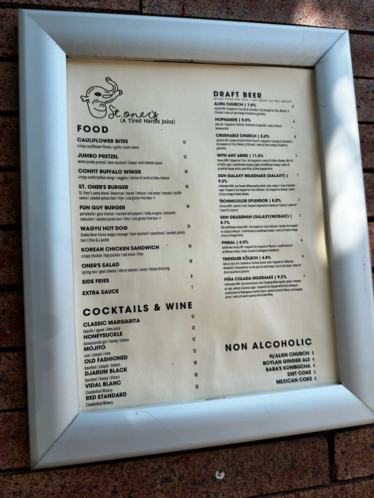 Menu at Stoner’s (A Tired Hands Joint) mounted on a brick wall. The menu is divided into four sections:

Food – Includes items like Cauliflower Bites, Jumbo Pretzel, Confit Buffalo Wings, St. Oner’s Burger, Fun Guy Burger, Wagyu Hot Dog, Korean Chicken Sandwich, Oner’s Salad, Side Fries, and Extra Sauce. Prices range from $2 to $14.

Cocktails & Wine – Offers drinks like Classic Margarita, Honeysuckle, Mojito, Old Fashioned, Djarum Black, Vidal Blanc, and Red Standard. Prices range from $10 to $14.

Draft Beer – Lists beers like Alien Church, HopHands, Crushable Church, With My Arms, DDH Galaxy Milkshake, Technicolor Splendor, Pizza Boy, Pineal, Tiredner Kölsch, and Piña Colada Milkshake with ABVs and brief descriptions. Prices mostly at $7.

Non Alcoholic – Includes N/Alien Church, Boylan Ginger Ale, Baba’s Kombucha, Diet Coke, and Mexican Coke, priced from $3 to $6.