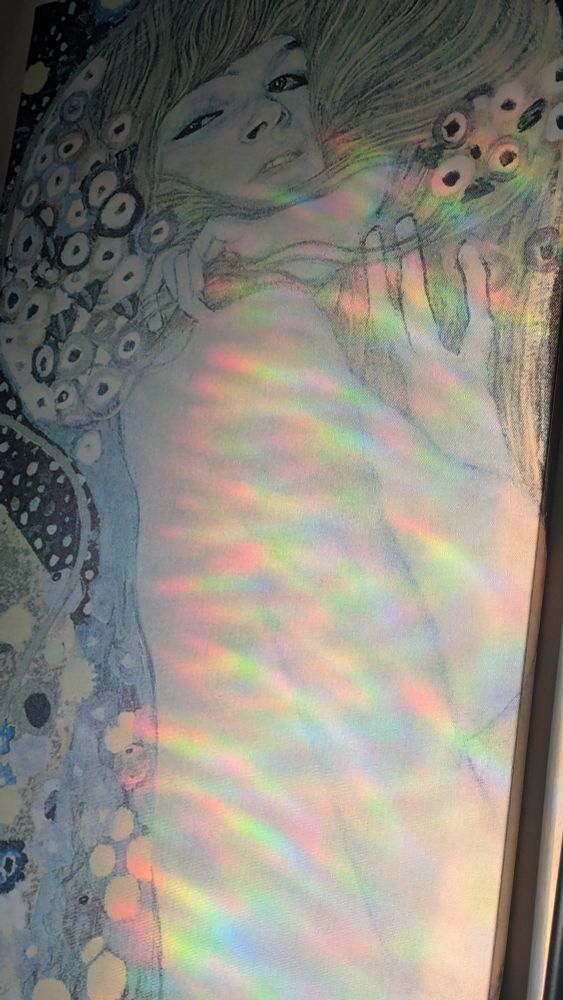 A myriad of rainbows are cast across a print of a part of Gustav Klimt's Water Snakes 2. The print features the side profile of a nude woman with her hands in her hair, surrounded by fish and flowers.
