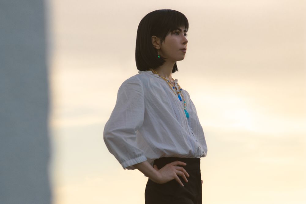 Cosplay photo of Howl Pendragon from the Ghibli film Howl's Moving Castle. I'm visible from the waist up, standing hands on my hips and looking into the distance against the pale sky, clouds illuminated yellow in the sunset. I'm wearing a white loose shirt and black pants, two necklaces with blue glass pendants and green earrings. 
