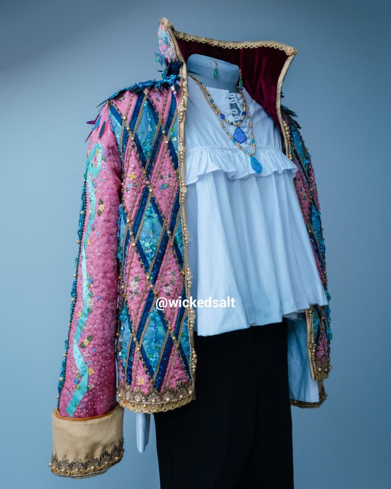 Photo of my Howl Pendragon cosplay on a sewing form, frontal three quarters view. White shirt and black pants with a pink and blue jacket thrown over the shoulders, like the Studio Ghibli design. The jacket is heavily beaded and covered in floss and ribbon embroidery. There are also two necklaces with blue pendants around the form's neck and a pair of green drop earrings pinned close by.