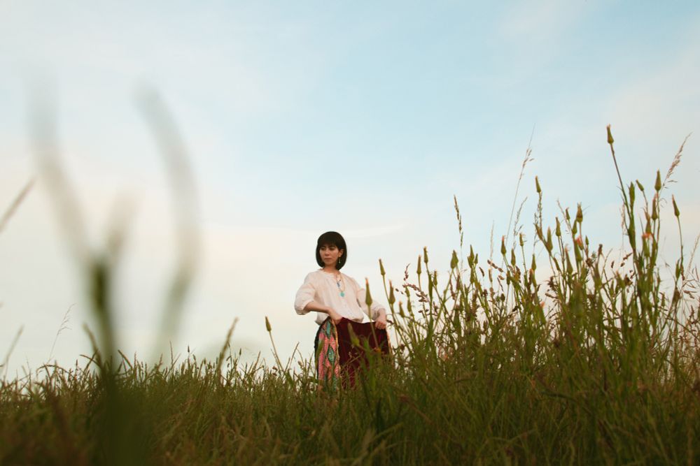 Cosplay photo of Howl Pendragon from the Ghibli film Howl's Moving Castle. I'm standing in tall green grass, holding the pink and blue jacket up in front of me. The sky is pale blue with white clouds. I'm wearing a white loose shirt and black pants.