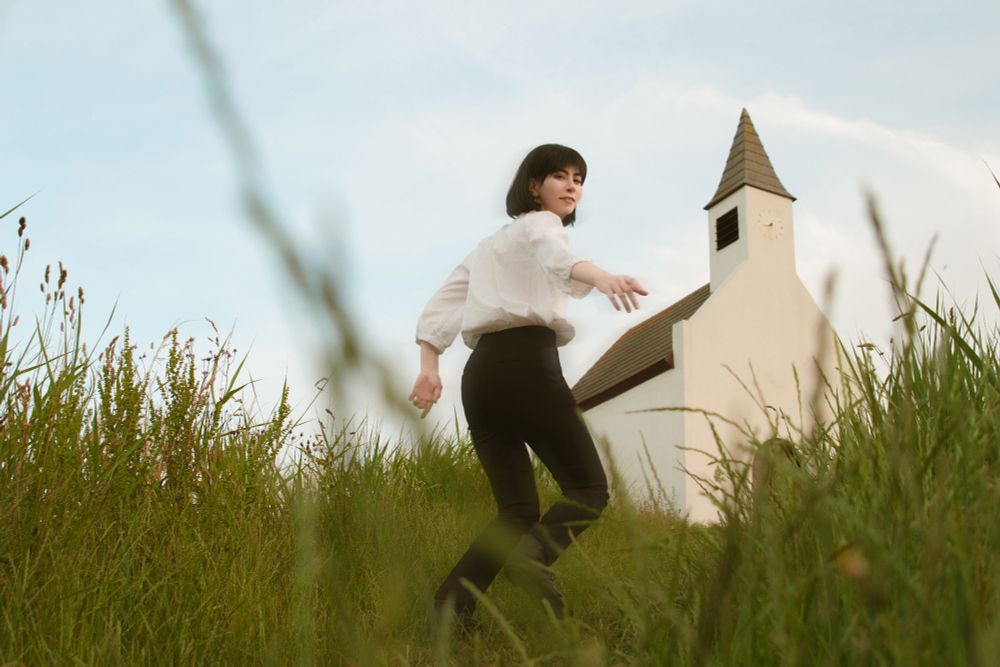 Cosplay photo of Howl Pendragon from the Ghibli film Howl's Moving Castle. I'm running up a hill of tall green grass and reaching behind me, hand extended to the viewer, the sky pale blue with white clouds and a white chapel on the hill. I'm wearing a white loose shirt and black pants.