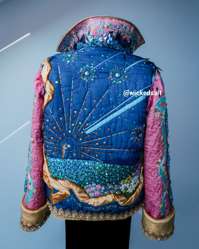 Photo of my Howl Pendragon cosplay on a sewing form, view from the back. Black pants pinned to the sewing form with a pink and blue jacket thrown over the shoulders, like the Studio Ghibli design. The jacket is heavily beaded and covered in floss and ribbon embroidery. The back of the jacket shows the scene from the Ghibli film as a young Howl standing on a meadow catches a falling star. There are other falling stars in the night sky. The meadow is completely beaded and framed in a golden velvet ribbon. Embroidered with golden floss beneath the golden ribbon reads: "You who swallowed a falling star, o' heartless man".