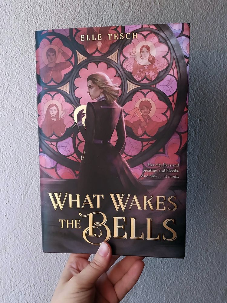 Photo of a hand holding the book What Wakes the Bells by Elle Tesch, cover illustration by Eleonor Piteira, featuring a young white woman with light brown hair, with her back towards the viewer, face in profile, in front of a circular gothic-style window with stained glass. There are 5 figures in the stained glass design. Smoke hides the bottom half of the woman while skulls carved in the stone beneath the window peek through.
