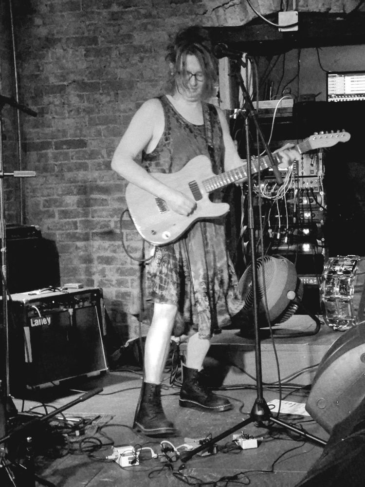 black and white shot of Selena - that's me - on the brick walled basement stage of Baba Yaga in Seattle's Pioneer Square — playing a home made butternut telecaster with one p90 in bridge position, not made by me by the way but I love it. I'm wearing a purple-green dress & black Docs & staring at me pedals as though I've never seen one. Rawk!