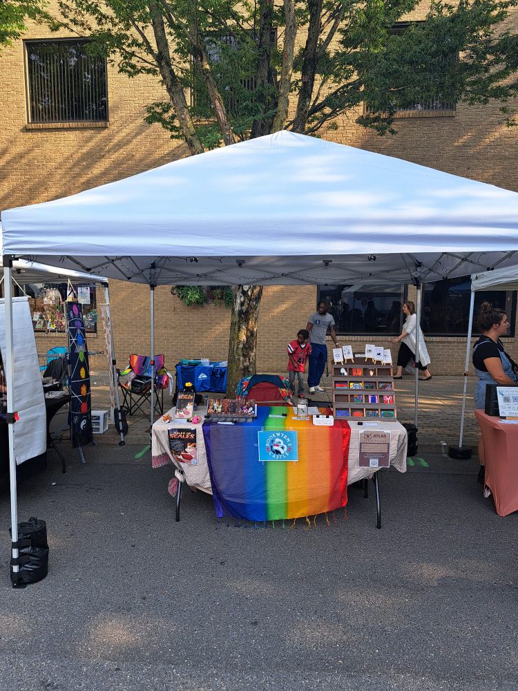 Picture of a booth setup with a white tent and rainbow table runner. There are books, bookmarks, paintings, and flash fiction booklets on the table
