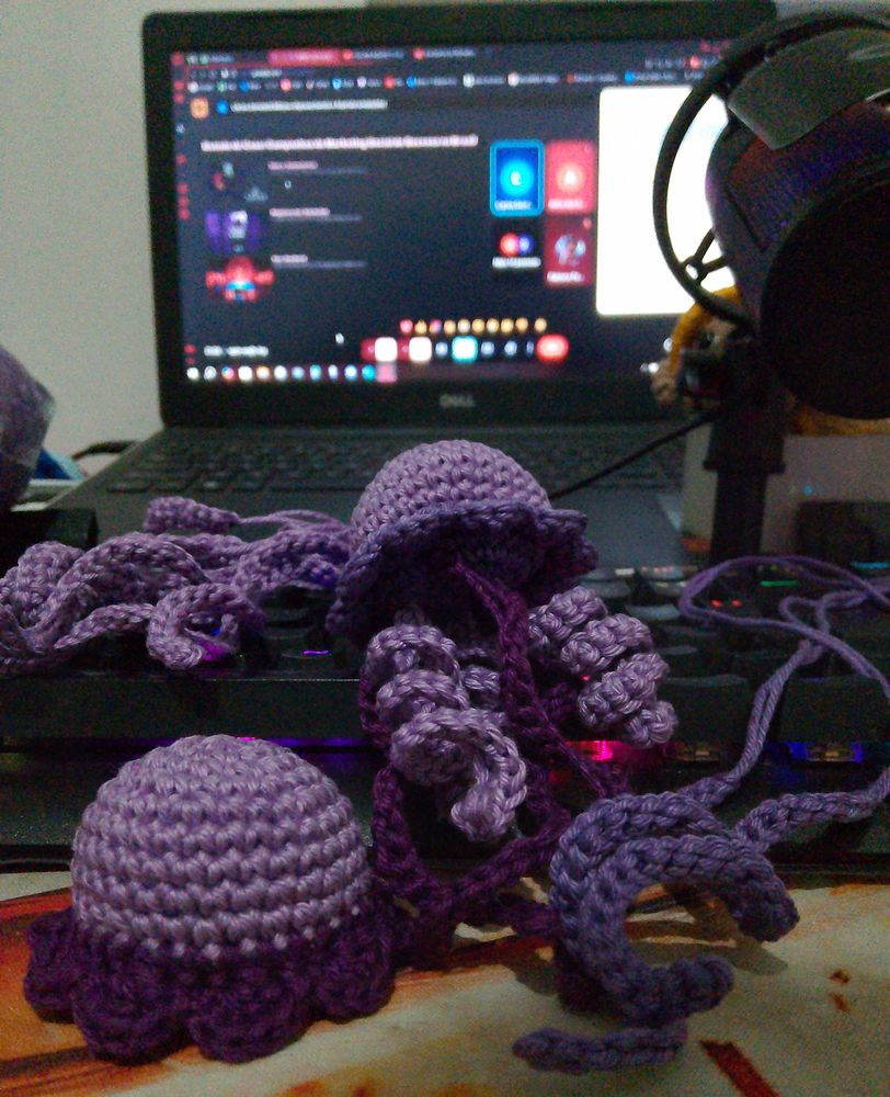 Chaveiros de água-viva em tonalidades de roxo sendo produzidos, pousados em cima de um teclado. 
Tela de notebook ao fundo mostrando apresentação em slide no Mett de um curso de Marketing pelo Senai.