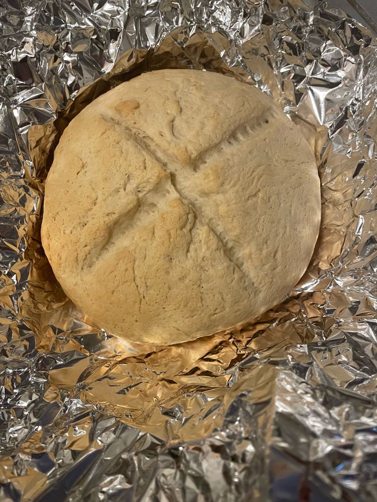 A round loaf of homemade bread with a cross scored into it. It's sitting on tin foil 