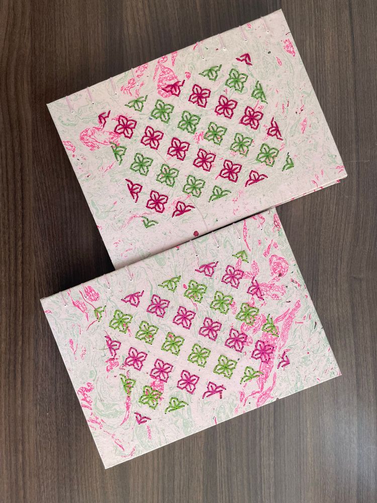 The photo contains two journals. The journals are handmade using coptic stitch binding. The cover of these journals have a pattern embroidered. The pattern is of a flower with four petals. Pink and green is used alternately for these flowers. 
