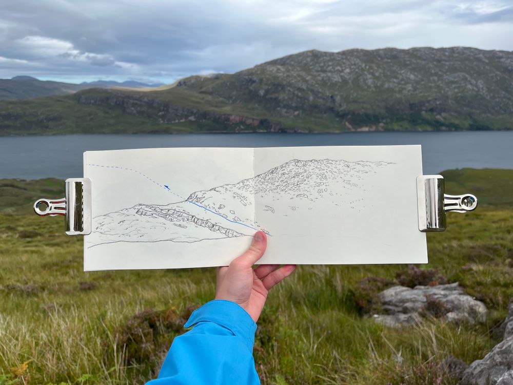 A hand holding a sketchbook with a pencil sketch of a mountainous landscape