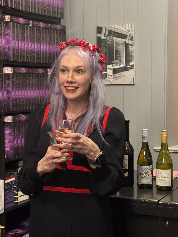 A K Benedict with lilac hair, red lips and in a long, black and red dress, holds a glass of fizz and grins 
