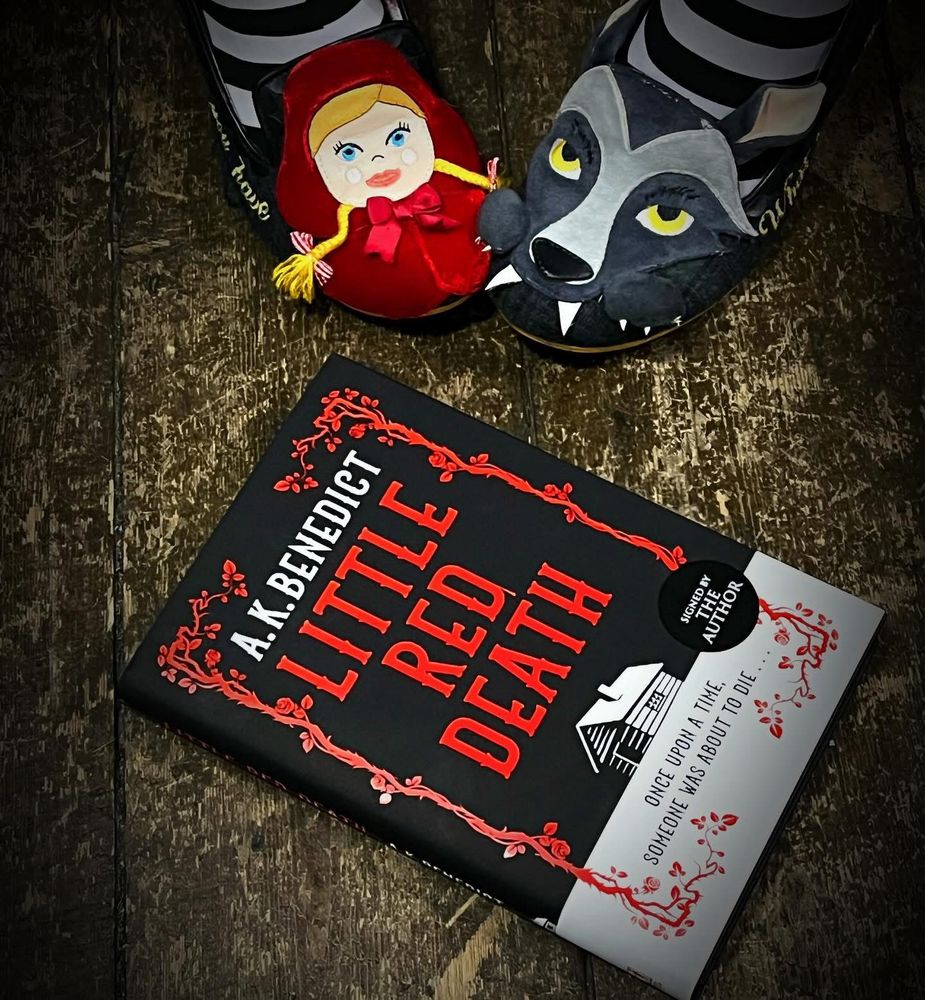 hardback book of Little Red Death by A K Benedict, on a wooden floor, next to a pair of shoes featuring Little Red Riding Hood and the big bad wolf 