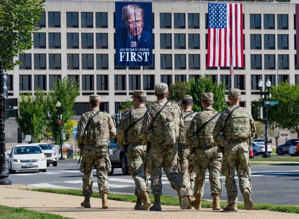 Une patrouille militaire armée et en treillis marche sur une allée à côté d'une route. A l'arrière plan, un grand bâtiment avec une grande affiche de Donald Trump et un drapeau américain qui sont affichés le long du mur sur à peu près trois étages