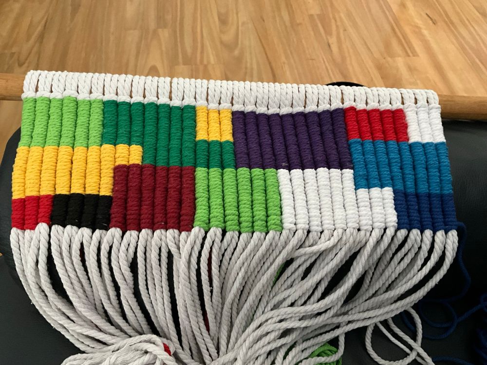 Unfinished macrame project. Wooden dowel holding white working cords. Cords of different colours are added to it creating a woven carpet style mat. The pattern is a collection of mixed squares and rectangles of different sizes in yellow,light green, dark green, white, light and dark blue and light and dark red.