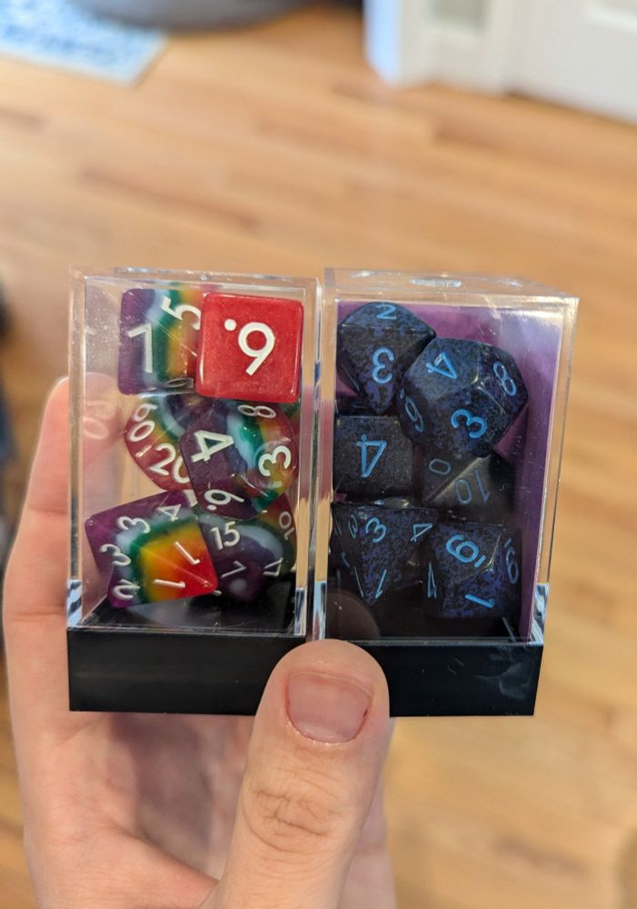 Hand holding two containers of dice sets. The set on the left is rainbow with white lettering and the set on the right is dark blue with light blue lettering. 