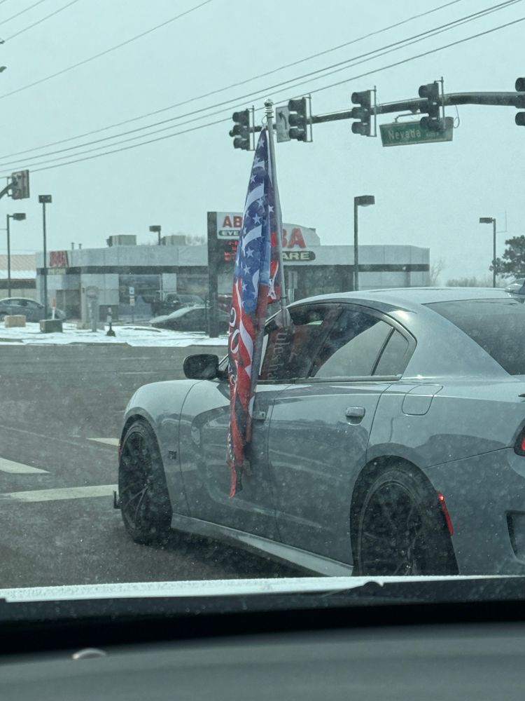 A muscle car with its window rolled down about 5 inches so that he can hold his Trump flag while driving down the street. 