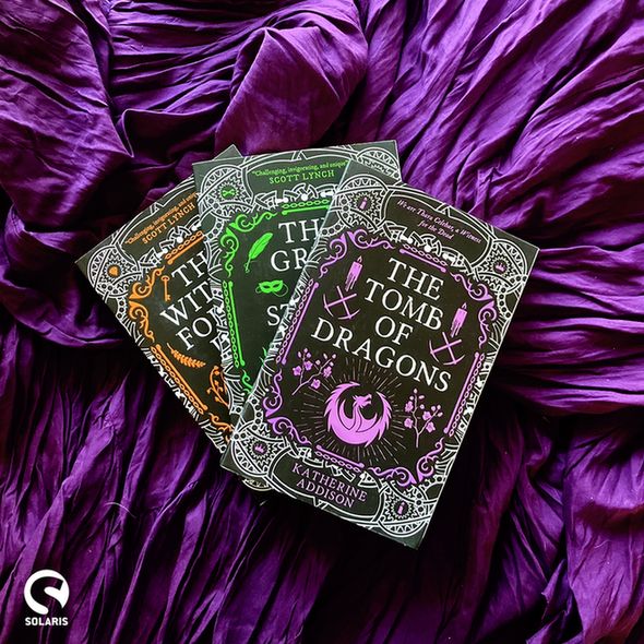Against a background of purple fabric, a fanned out stack of the three CEMETERIES OF AMALO series: THE WITNESS FOR THE DEAD, THE GRIEF OF STONES and THE TOMB OF DRAGONS by Katherine Addison. The white Solaris logo is in the bottom left corner.