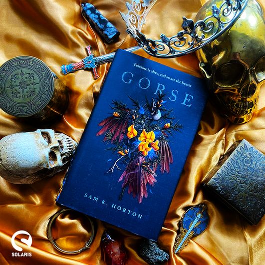Against a gold silk background, surrounded by skulls, crowns, swords, gold objects and crystals: a hardback copy of GORSE by Sam K. Horton. The white Solaris logo is in the bottom left corner.