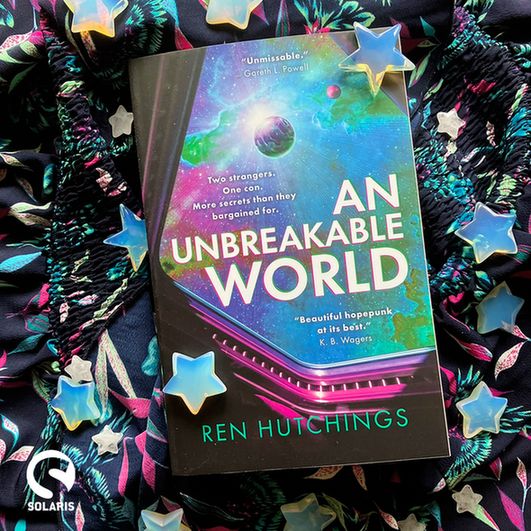 Against a background of navy, teal and pink patterned fabric dotted with stars, a flatlaid paperback copy of AN BREAKABLE WORLD by Ren Hutchings. The white Solaris logo is in the bottom left corner.