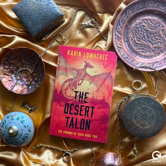 Against a gold silk background, a flatlaid novella paperback -  THE DESERT TALON by Karin Lowachee. The book is surrounded by gold and copper objects. The white Solaris nova logo is in the bottom right corner.