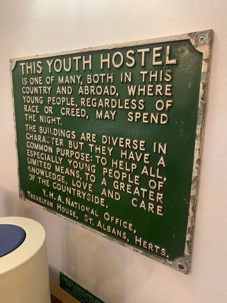 Old fashioned green metal plaque reading, “This youth hostel is one of many, both in this country and abroad, where young people, regardless of race or creed, may spend the night.  The buildings are diverse in character but they have a common purpose:  to help all, especially young people of limited means, to a greater knowledge, love and care of the countryside.  YHA national office, Trevelyan House, St Albans, Herts.”