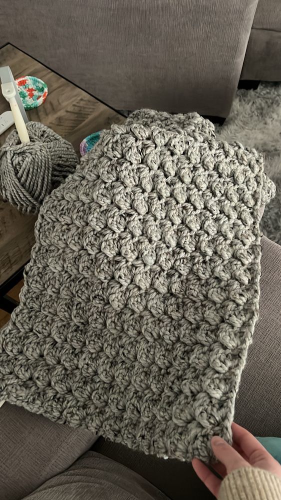 Picture showing Monica’s progress on a blanket she is crocheting, with a ball of yarn on a yarn holder on the table in the top right corner. The blanket, which is roughly a foot wide, is made with a thick, light grey wool-type yarn with specks of black and brown. The stitches in the blanket look like small bubbles.