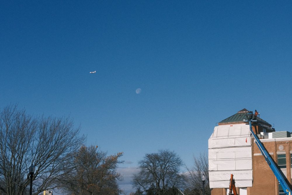 Morning sky with faint moon, plane passing by, construction workers on the top of a building, bare trees all around