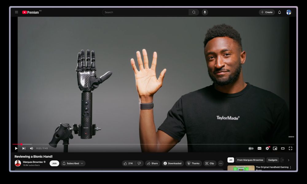 Marques Brownlee, a tech YouTuber, stands next to a robotic bionic hand mounted on a stand. Both he and the bionic hand are holding their palms up, mirroring each other. Marques is smiling and wearing a black TaylorMade t-shirt. The scene is from a YouTube video titled “Reviewing a Bionic Hand!”

Disclaimer: Alt text is AI generated.