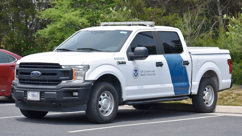 This is a similar vehicle to the last image except instead of an SUV its a white truck and instead of a green stripe it's a blue stripe. It still has police lights, and identifying writing on the side