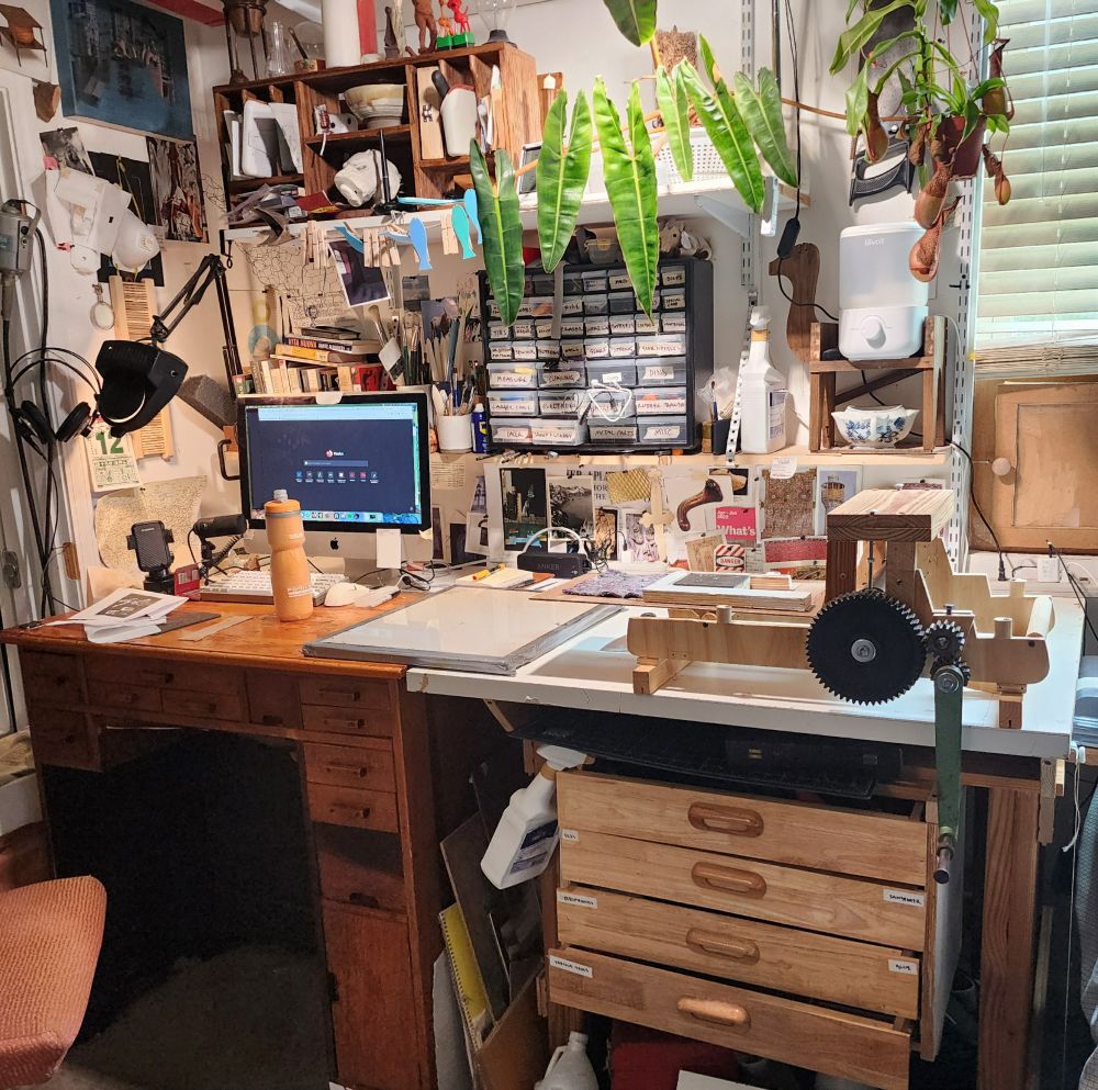 An image of my workspace with my computer, a large palette, and a printing press on my desk. There are lots of little things and drawers and images on the walls