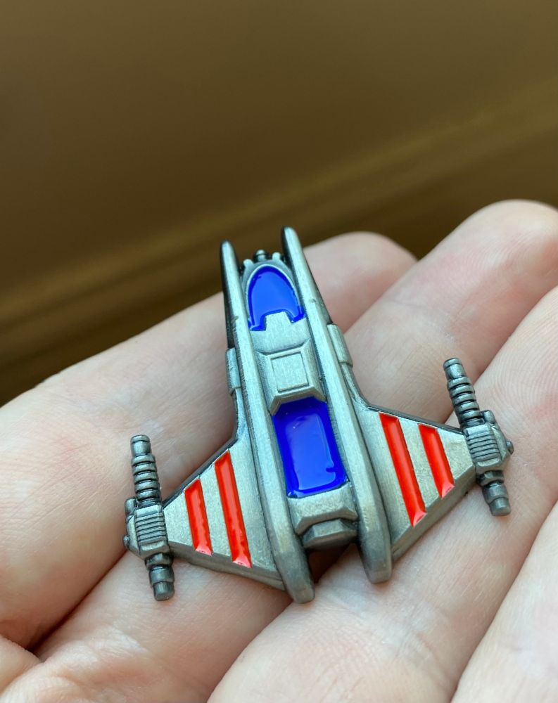 A person’s palm holding a pin of the Chmmr Avatar, a spaceship from Free Stars: The Ur-Quan Masters and Children of Infinity. 