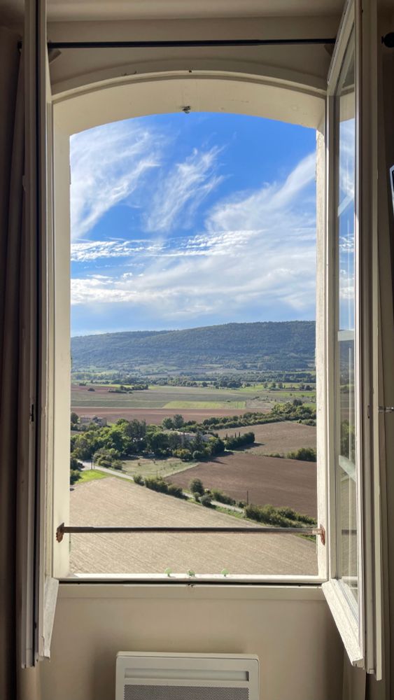 The photo shows the view through a window of a Provençal landscape on a sunny day. Lavender fields are in the foreground and tree-covered hills in the background.