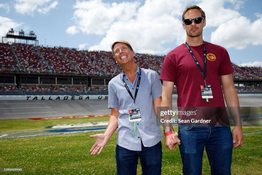 Jack McBrayer and Alexander Skarsgard holding hands while standing on the field in a stadium. McBrayer is smiling or in the middle of laughing while Skarsgard is looking at the camera. They are adorable.