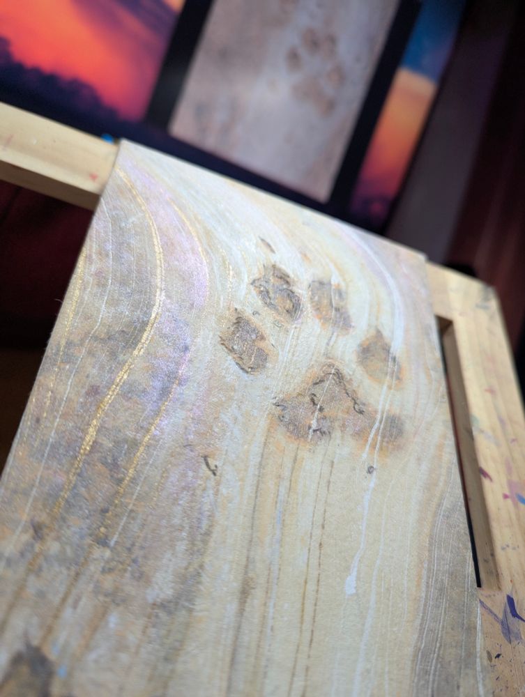 A low angle photo of a work in progress painting of a coyote footprint in sandy earth, with shimmers of gold and violet where the light hits it. The reference photo for the painting is on an out of focus computer monitor in the background. 