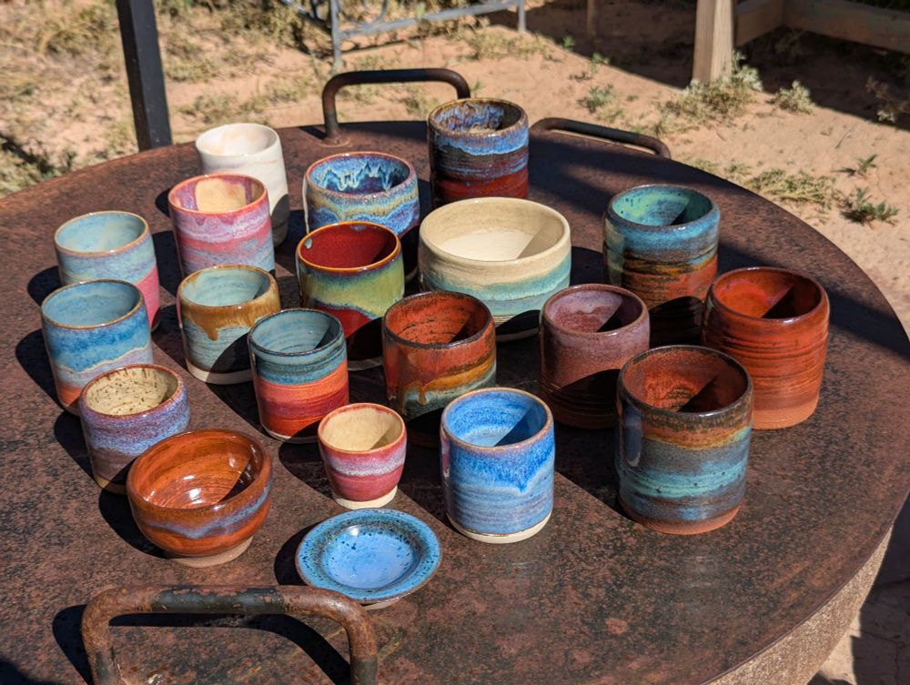 20 small ceramics pieces, mostly cups, arranged on a fire ring lid. The glazes on the ceramics range from warm orange earth tones, various shades of turquoise, some reds, blues, and pinks. 