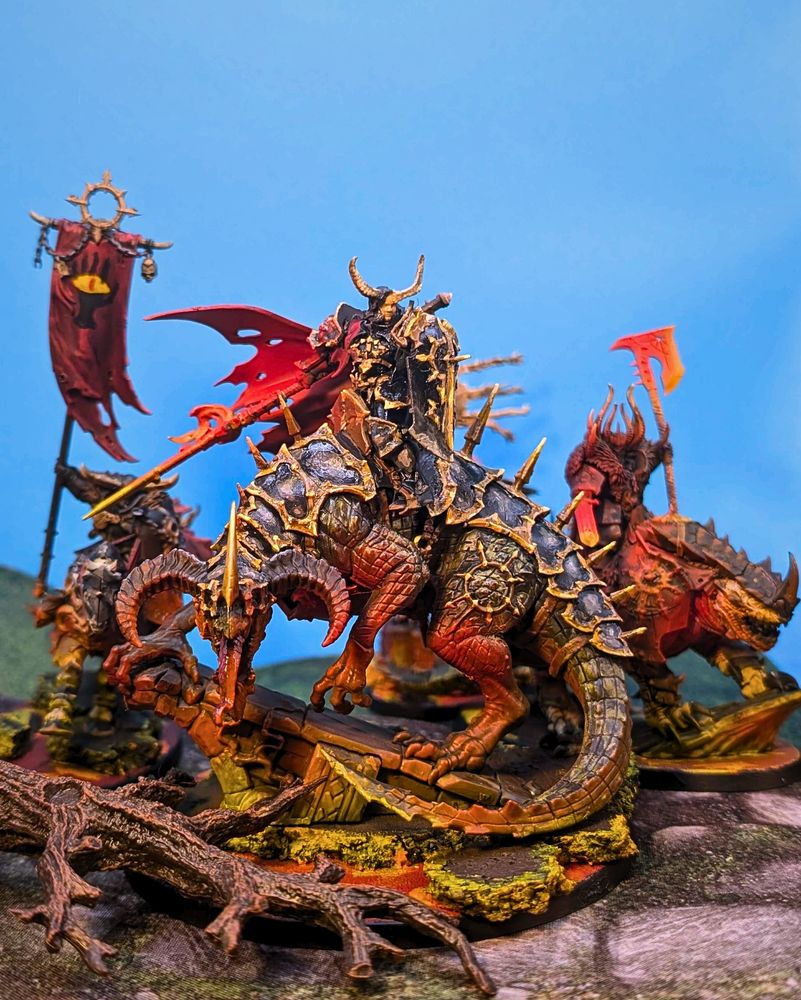 A group of mounted chaos warriors charging through a forest path. The leader rides a massive armored reptilian beast, and a red banner featuring a black claw grasping a yellow eye flies behind her.