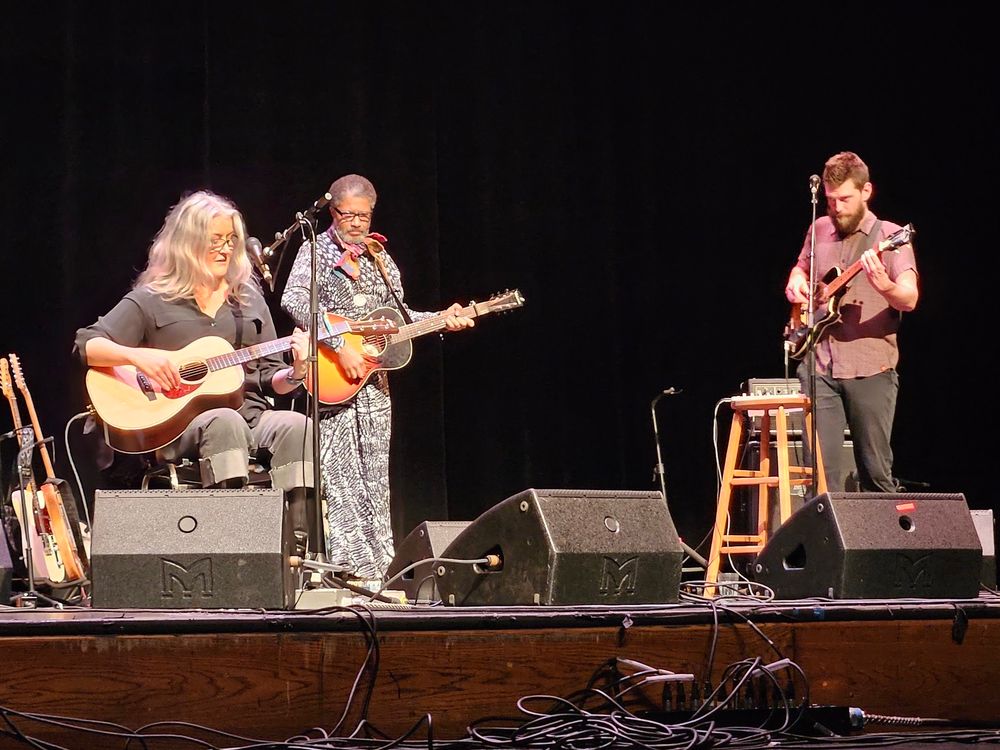 Paula Cole seated, playing guitar, with two band mates behind her.