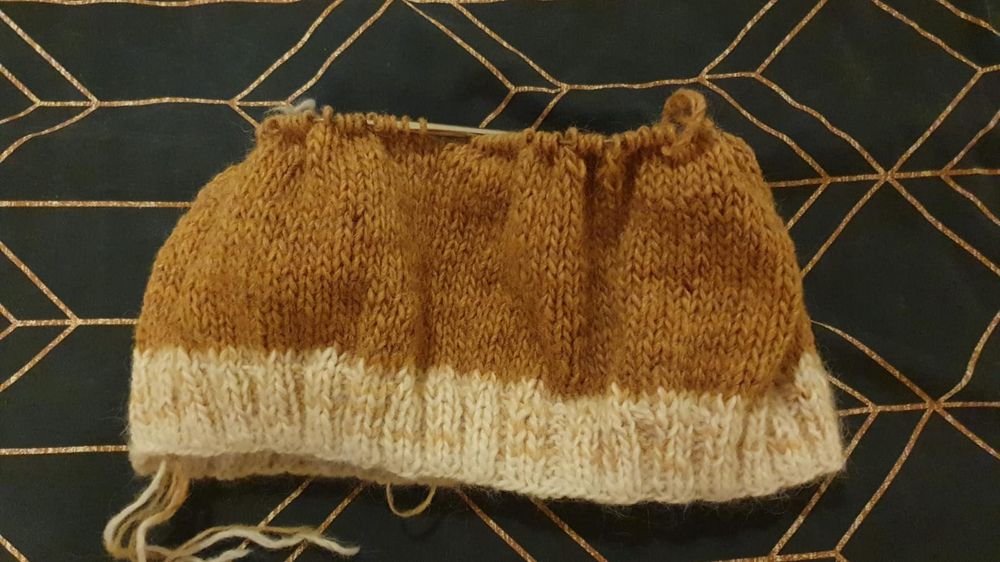 A knitted jumper in progress on a dark blue background with geometric pattern of intersecting kite-like golden or copper-colored lines. 10 rows of 2k2p white and yellow hem (a mix of white Álafosslopi and bright yellow plötulopi) and about 25 rows of knit stitches in rusty yellow (Álafosslopi standard yellow) on 6mm round needles. 
