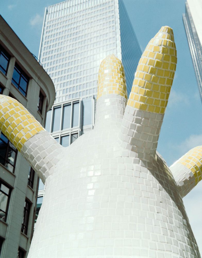 Tall sculpture in the City of London. Covered in small yellow and white square tiles. An office building is in the background.