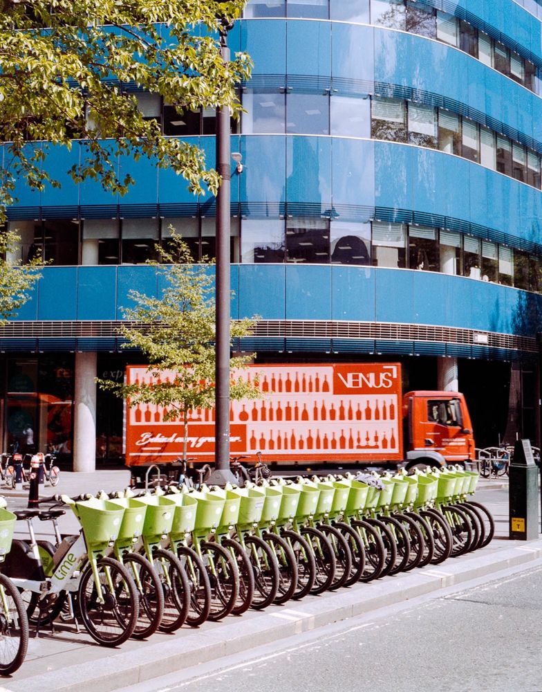 City of London street with a row of Lime bikes. A blue office building is in the background and a red truck drives by.
