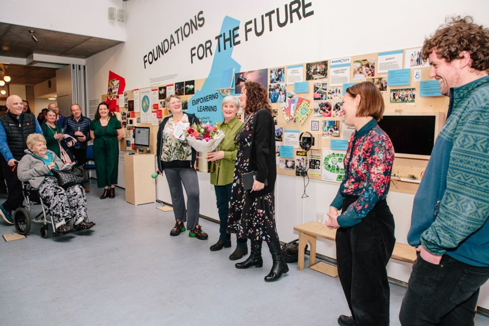 A lively gathering at the Foundations for the Future exhibition, with a group celebrating in front of a wall showcasing community projects and learning initiatives.