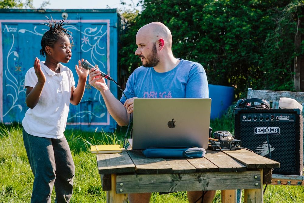 Child speaking into a microphone held by an adult with a laptop and amp outdoors.